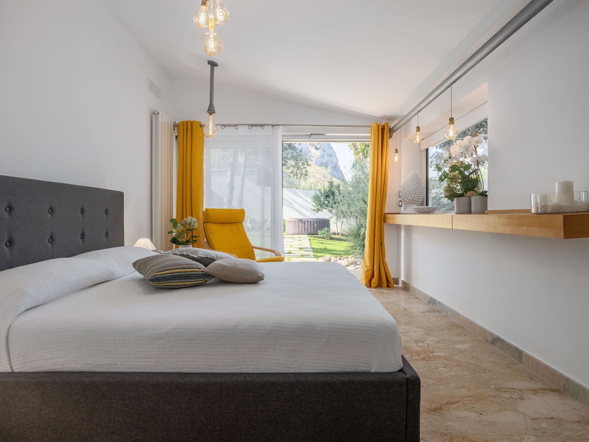 Una camera da letto con un letto, una sedia, una finestra con tende e uno scaffale con oggetti decorativi.