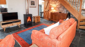 A cozy living room with a fireplace, a sofa, a TV, and a staircase leading to a stone wall. - Post Office Cottage (Goodwick)