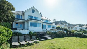 A large, modern house with a spacious front yard, a set of wooden chairs, and a stone staircase leading to the entrance. - Treverden (Wadebridge)