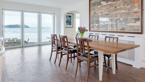 A dining area with a wooden table and chairs, a large abstract painting, and a view of the ocean through glass doors. - Treverden (Wadebridge)