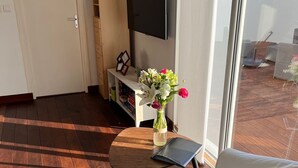 Un salón con un sofá blanco, una mesa de centro de madera con un jarrón de flores, un televisor en la pared y una estantería.