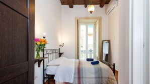 Quarto com cama, mesa de cabeceira com um vaso de flores, espelho e porta que dá para outro cômodo.