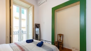 Quarto de hotel com cama, uma cadeira, uma mesinha, janela com vista e porta com moldura verde.