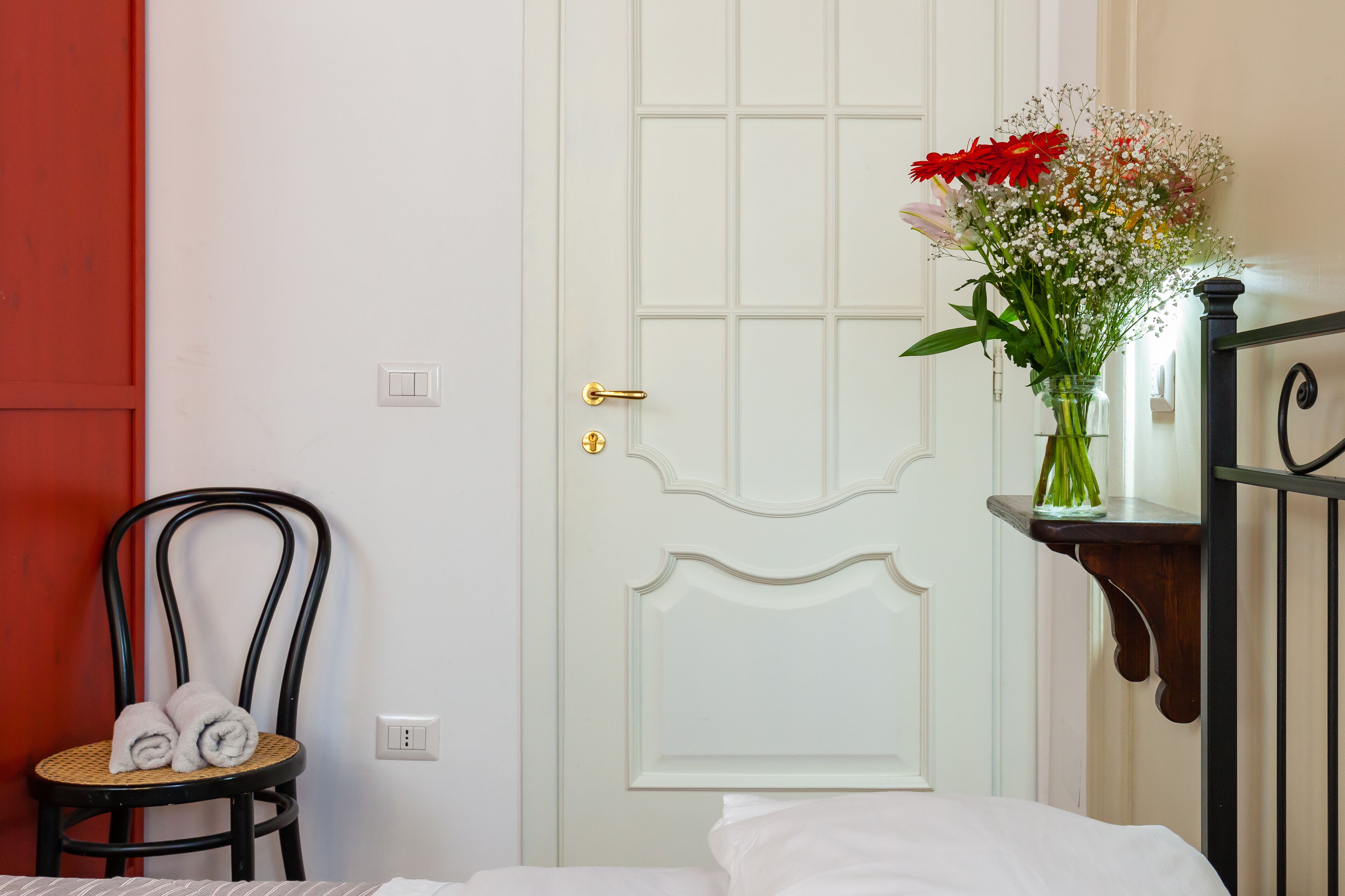 Ein Zimmer mit einem Bett, einem Sessel, Handtüchern, einer weißen Tür und einer Vase mit Blumen.