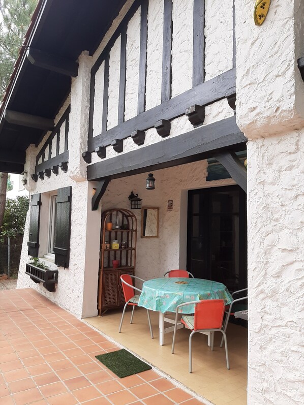 Un bâtiment à l’extérieur blanc et aux poutres en bois foncé, une table recouverte d’une nappe verte et des chaises rouges.
