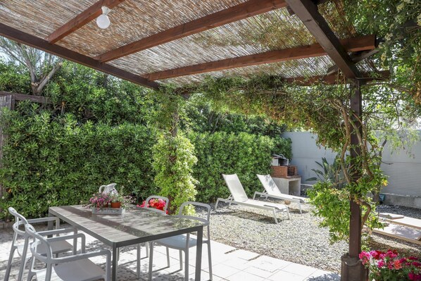 Relax in a serene outdoor space surrounded by lush greenery and a pergola, perfect for dining or unwinding with a view of the adjacent brick wall.