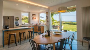 Een moderne keuken met een groot schiereiland, barstoelen en een eettafel met stoelen. Grote ramen bieden uitzicht op het omliggende landschap.