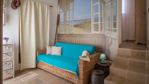 Un sofá de mimbre con funda color turquesa, una mesita con una lámpara y vista al mar a través de una ventana.