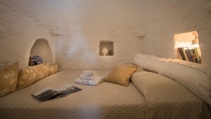Una habitación acogedora y bien iluminada con un sofá, cojines, una mesita con una revista y una estantería con libros y objetos decorativos.