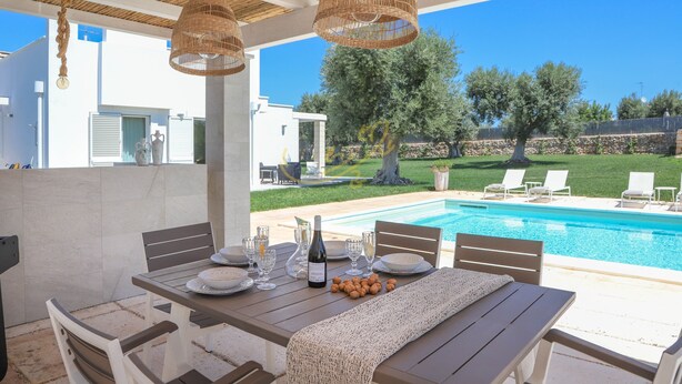 An outdoor dining area with a pool view, wicker hanging lights, and a table set for a meal. - TD Villa Patrizia Pool Alfresco Kitchen (Monopoli)