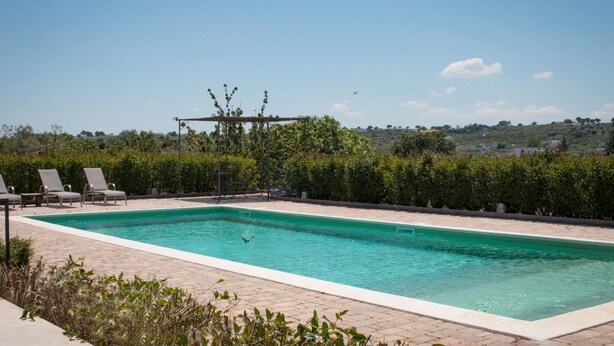 A rectangular swimming pool with a pergola and lounge chairs, surrounded by hedges and a view of a hilly landscape.
