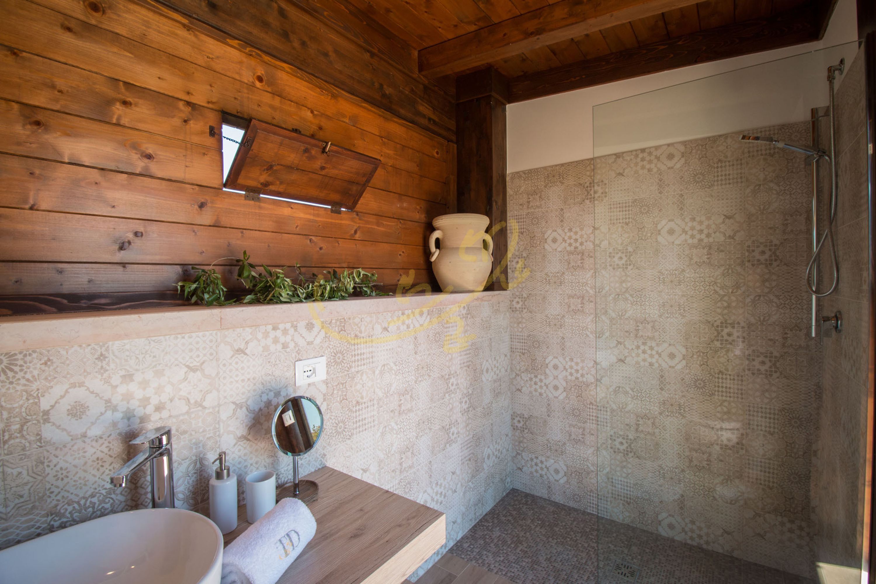 Un baño con techo de madera, un lavabo con espejo, ducha con azulejos estampados y un florero en la repisa.