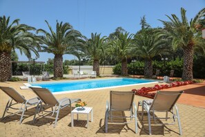 Area piscina con lettini e tavolo, circondata da palme.