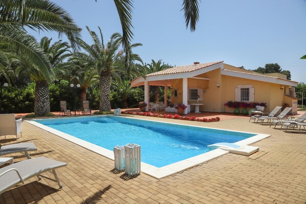 Una piscina con lettini, un'area salotto coperta e palme.