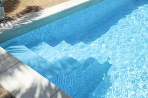 Una piscina azzurra e trasparente con scalini per entrarvi, circondata da un solarium piastrellato.