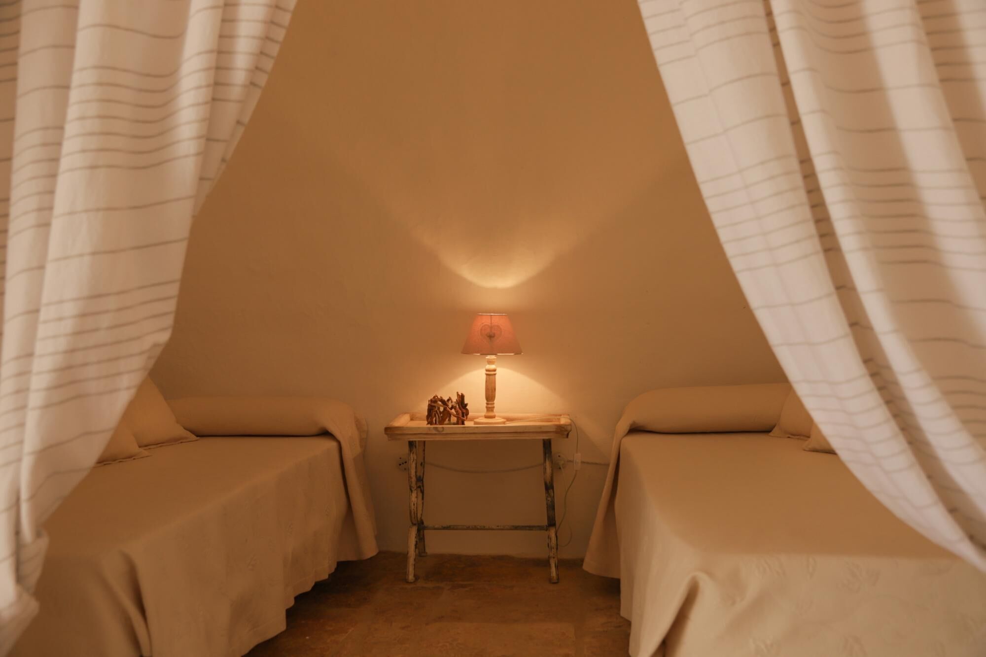 A cozy, triangular room with two beds, a small table with a lamp, and a wall-mounted shelf.