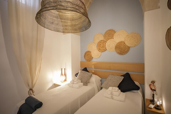 Une chambre d’hôtel avec deux lits, une tête de lit en osier, une décoration murale composée de cercles en osier, et une table de chevet avec une lampe et un objet décoratif.