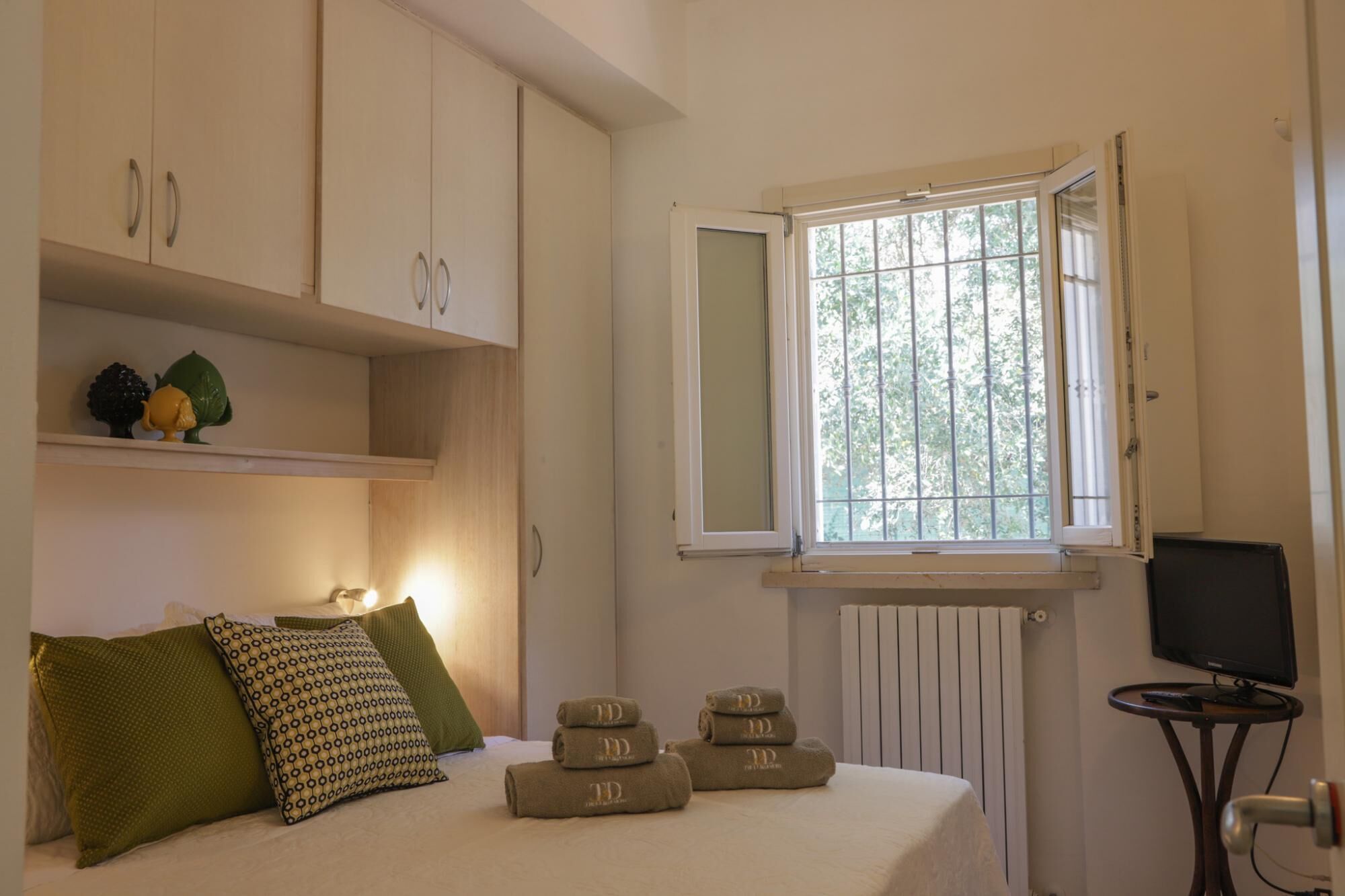 A bedroom with a bed, pillows, a window, a radiator, a television, and a small table.