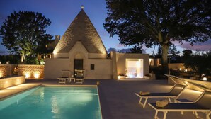 Une piscine avec des chaises longues, un bâtiment en pierre doté d’un toit conique et un arbre.