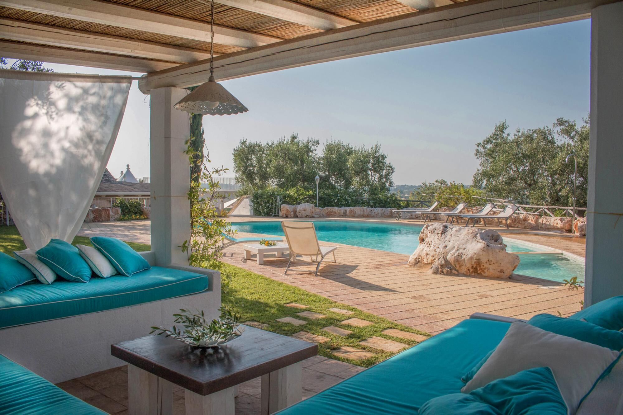 A covered outdoor seating area with a pool, lounge chairs, and a view of the surrounding landscape.