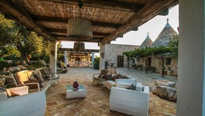 An outdoor patio with stone flooring, wicker furniture, and a pergola with a hanging light fixture. - TD Trulli di Montaltino With Stunning Huge Pool (Polignano a Mare)