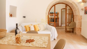 A cozy living room with a white sofa, a wooden coffee table with snacks, a bottle of wine, and a wicker chair. An arched doorway leads to a kitchen area. - TD Trulli Bruco Large Pool for Family Fun (Monopoli)