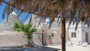 A patio area with a table set for dining, surrounded by white chairs, and a stone building with conical roofs in the background. - TD Trulli Bruco Large Pool for Family Fun (Monopoli)