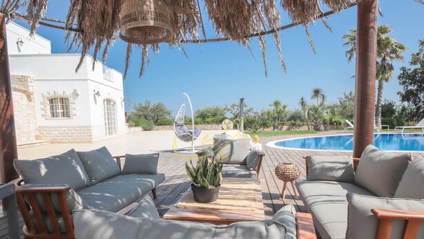 An outdoor seating area with a pool, wooden deck, and a white building in the background. - TD Trulli Bruco Large Pool for Family Fun (Monopoli)