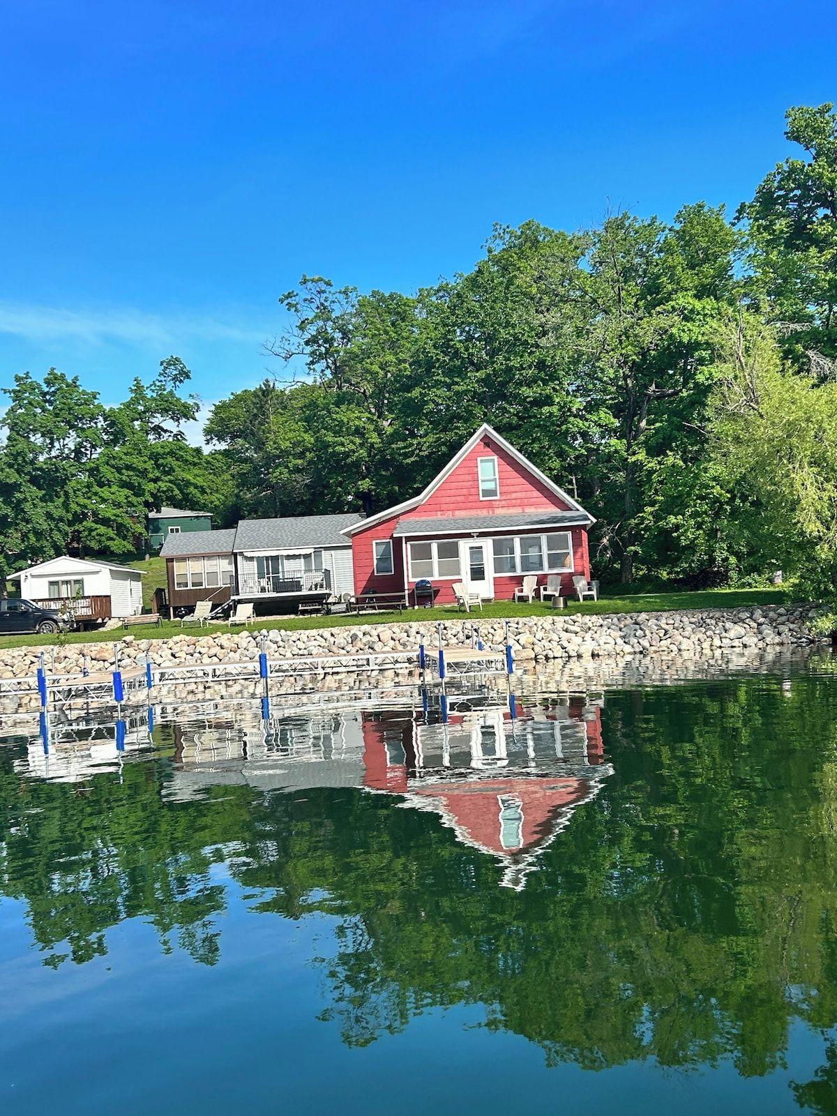 A red house with a stone wall and dock, surrounded by trees and calm water.