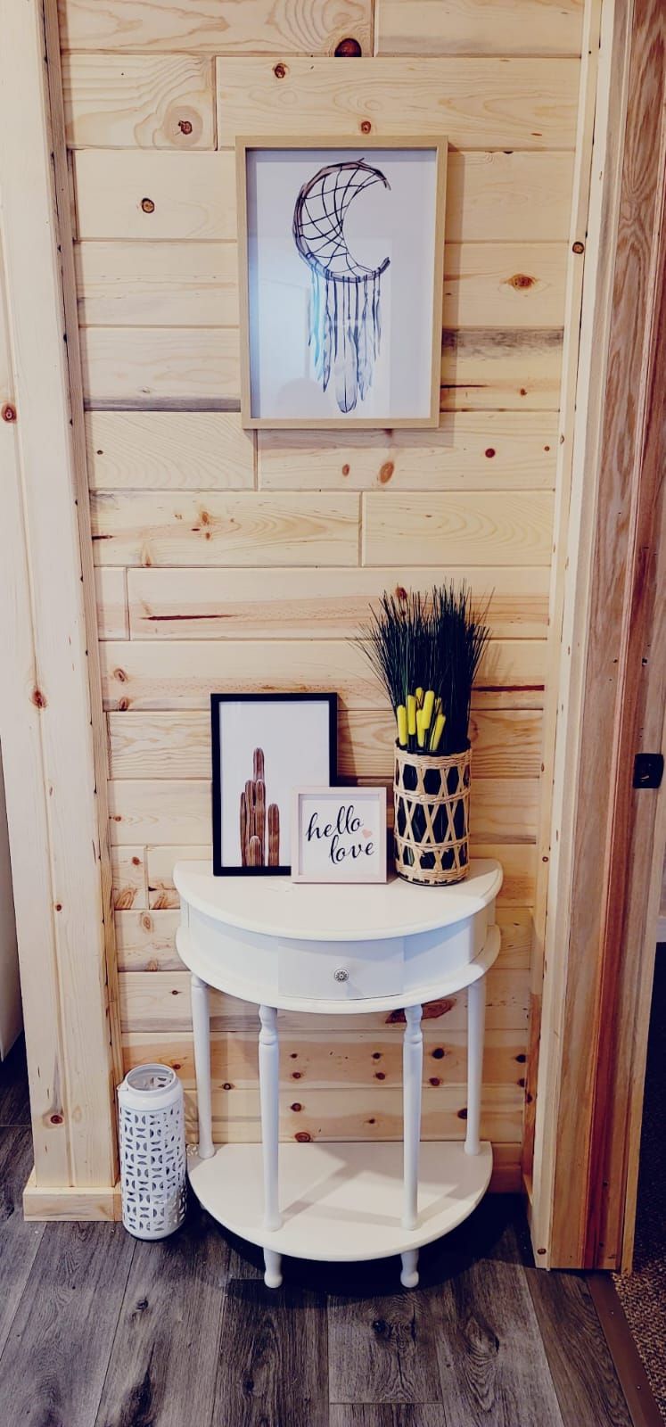 Un mur en bois orné d’un tableau encadré, une petite table blanche avec une pancarte indiquant « bonjour », et un vase décoratif contenant des fleurs jaunes.