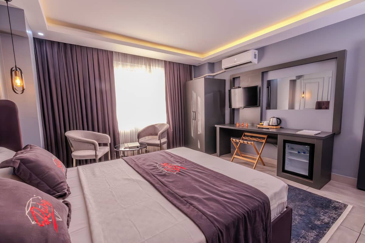 A modern hotel room with a large bed, a desk, a chair, a TV, and a minibar.