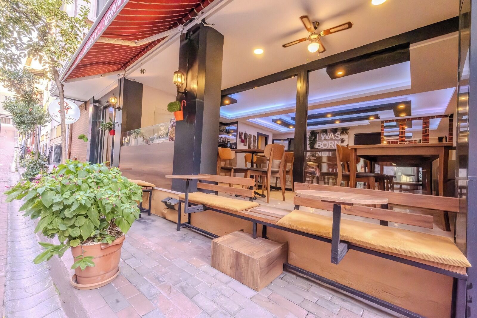 Un espacio exterior con mesas y bancos de madera, una planta en maceta y una fachada con un letrero.