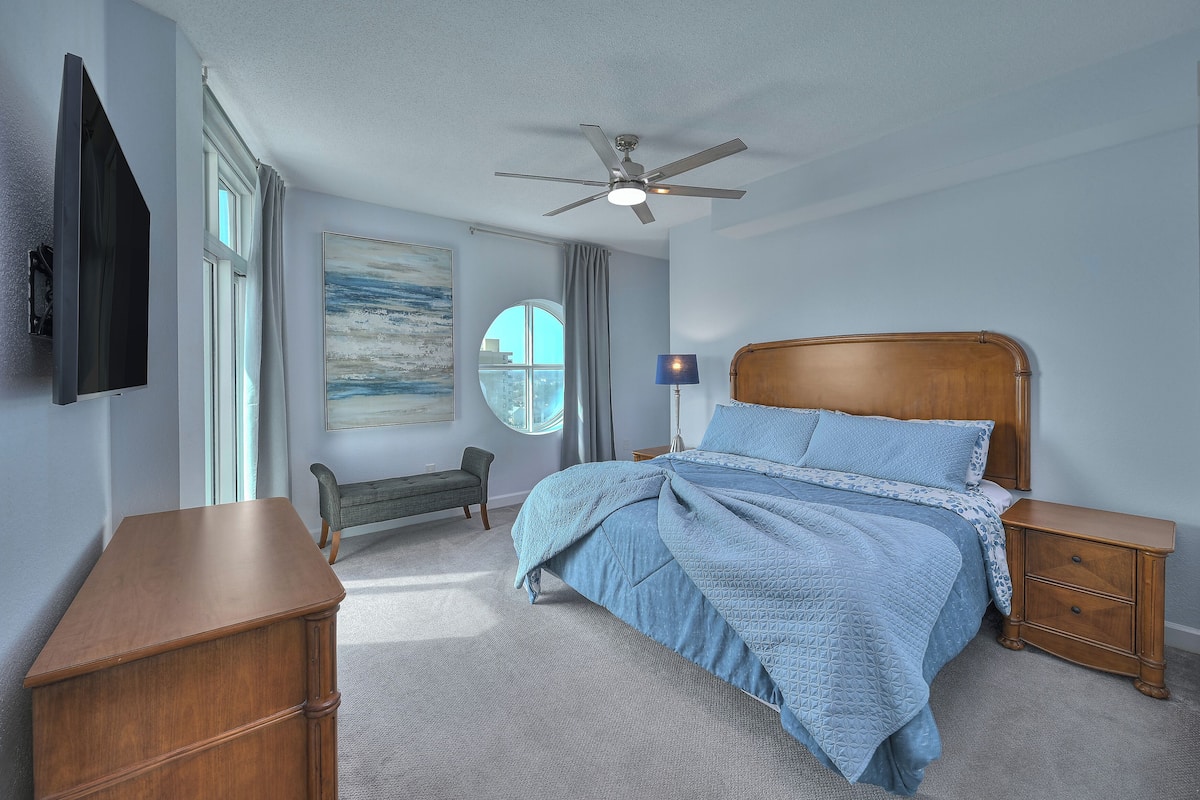 A bedroom with a bed, a desk, a chair, a nightstand, a lamp, a ceiling fan, and a window with curtains.