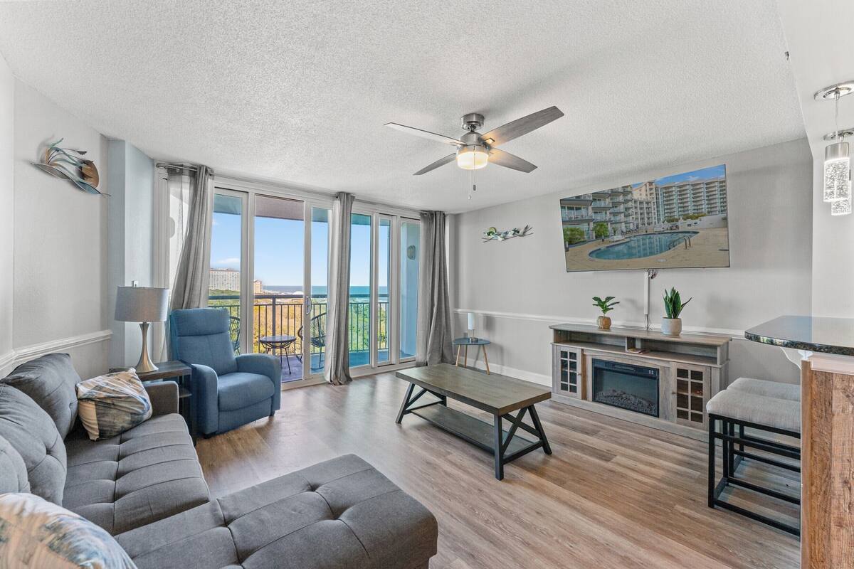 A modern living room with a sofa, armchair, coffee table, fireplace, and a balcony with a view.