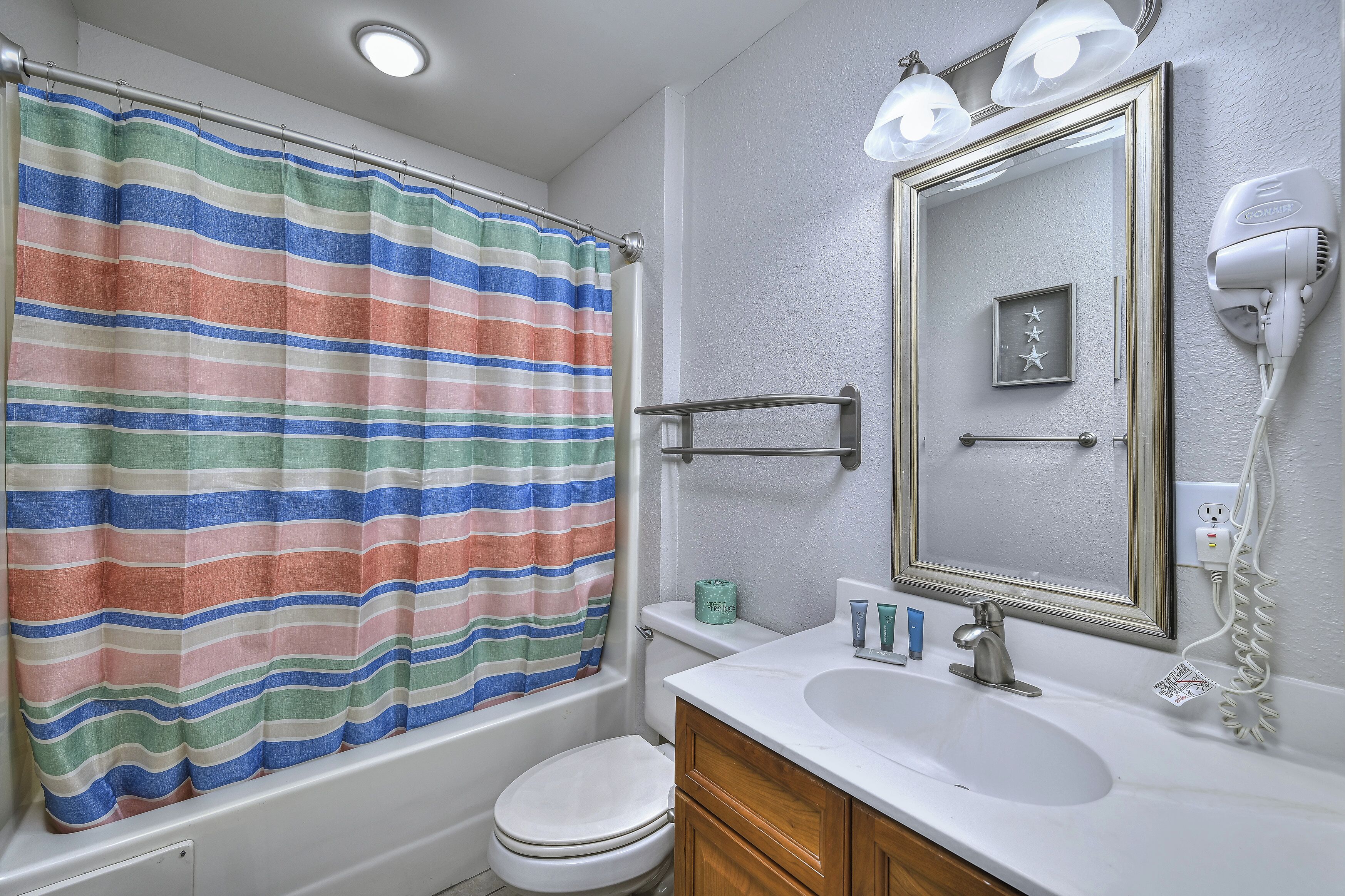 A bathroom with a striped shower curtain, a white toilet, a sink with a mirror, and a hairdryer on the wall.