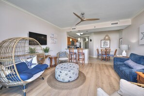 A living room with a blue sofa, a wicker chair, a round ottoman, and a dining area with a table and chairs. - Horizon at 77th by Elliott Beach Rentals (Myrtle Beach)