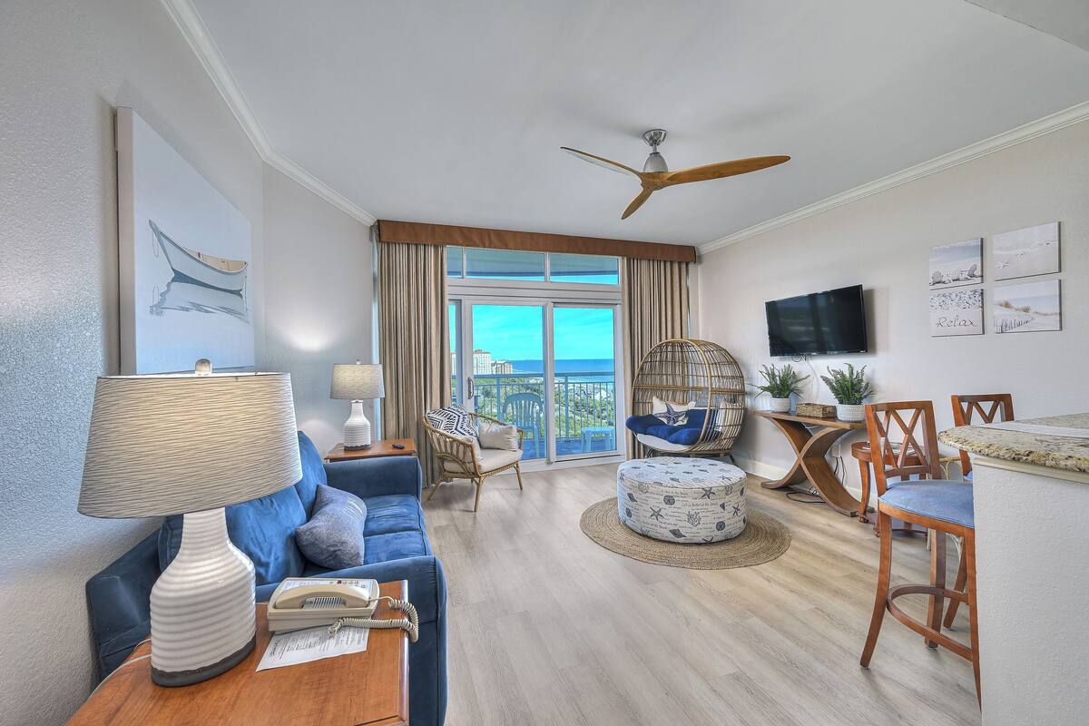 A spacious living area with a sofa, a coffee table, a dining table with chairs, a ceiling fan, and a large window with a view of the city.