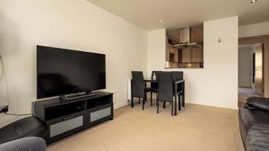 A modern living room with a flat-screen TV, a dining table with chairs, a sofa, and a kitchen area in the background.
