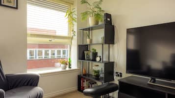Une pièce avec un canapé noir, un téléviseur, une bibliothèque avec des plantes et une fenêtre avec des stores.