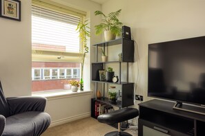 A room with a black sofa, a TV, a bookshelf with plants, and a window with blinds. - Sleek 2BDR City Stay in the Heart of Edinburgh (Edinburgh)