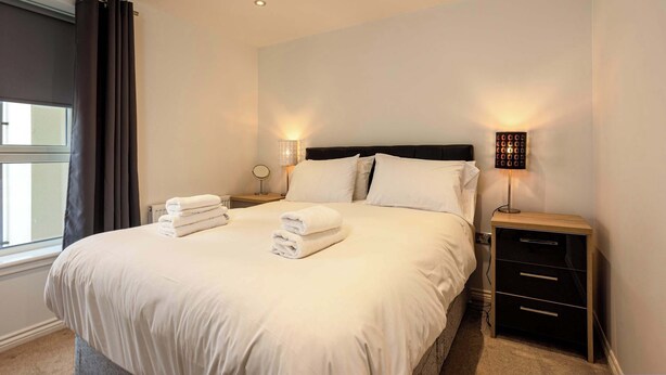 A neatly made bed with white linens, two bedside lamps, a nightstand with drawers, and a window with curtains.