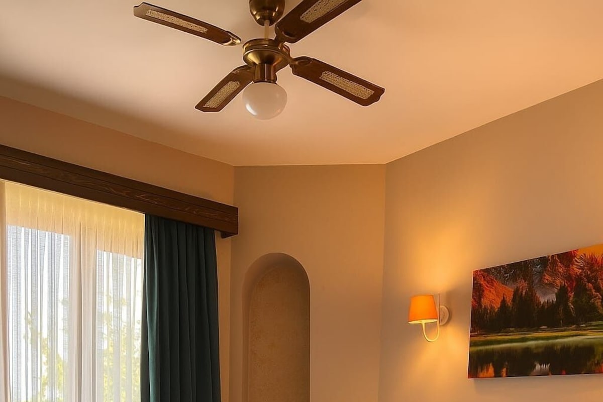A neatly made bed with white linens and towels, a ceiling fan, a painting of a landscape, and a chair in a room with beige walls and a wooden floor.