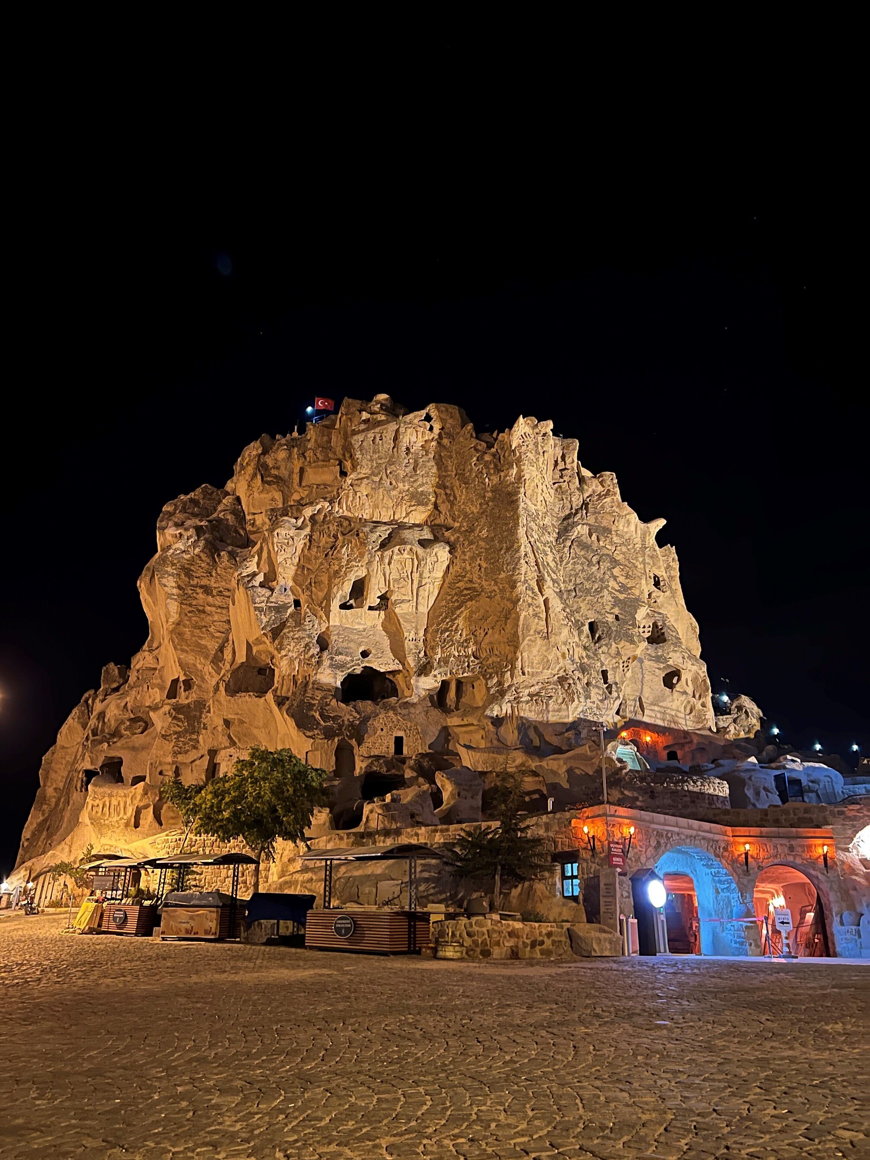 A large rock formation with carved structures and a flag, surrounded by a paved area with a building entrance.