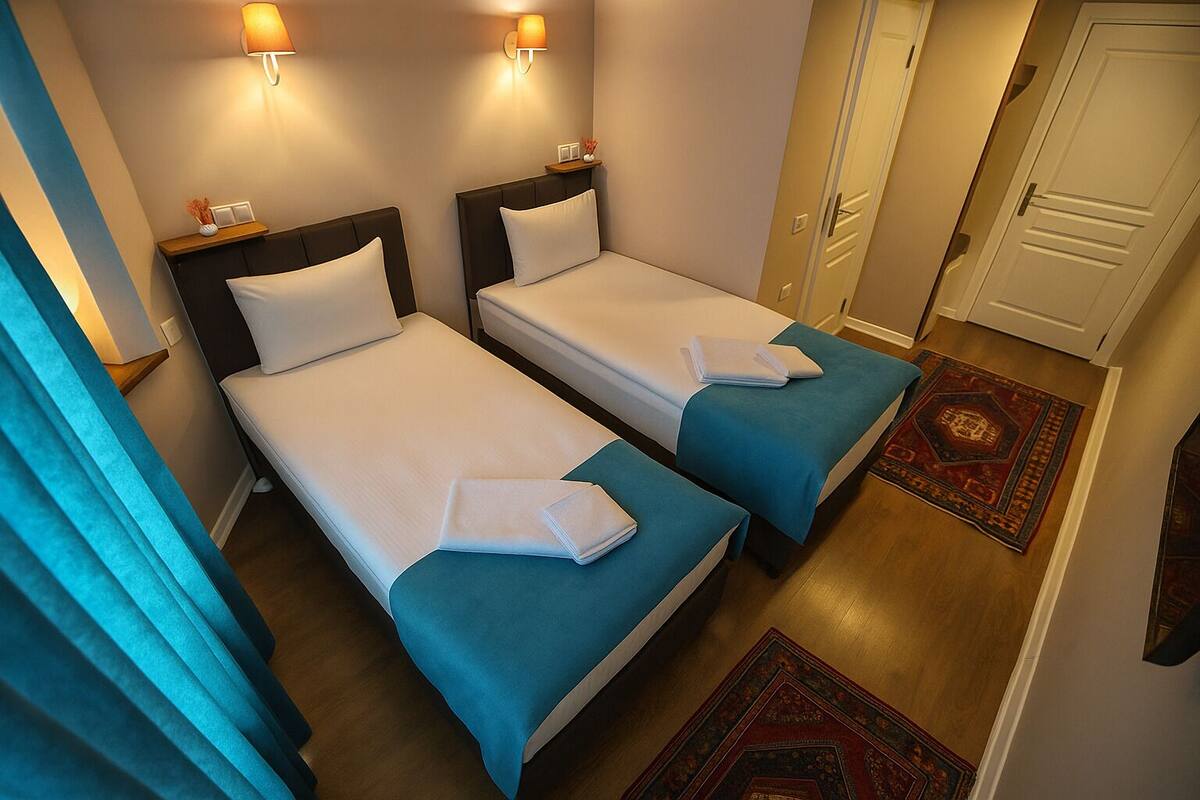 A hotel room with two single beds, each with a teal bedspread and white pillows. There are wall-mounted lamps on either side of the beds, and a wooden nightstand with a vase of flowers. The room has a wooden floor and a patterned rug near the door.