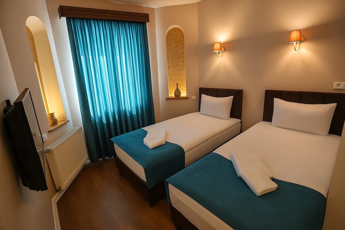 A hotel room with two beds, a window with curtains, a wall-mounted lamp, and a radiator.