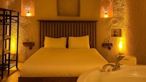A room with a bed, a wooden headboard, two wall-mounted lights, a large window with a view, and a bathtub. - Grand Uçhisar Hotel (Nevsehir)