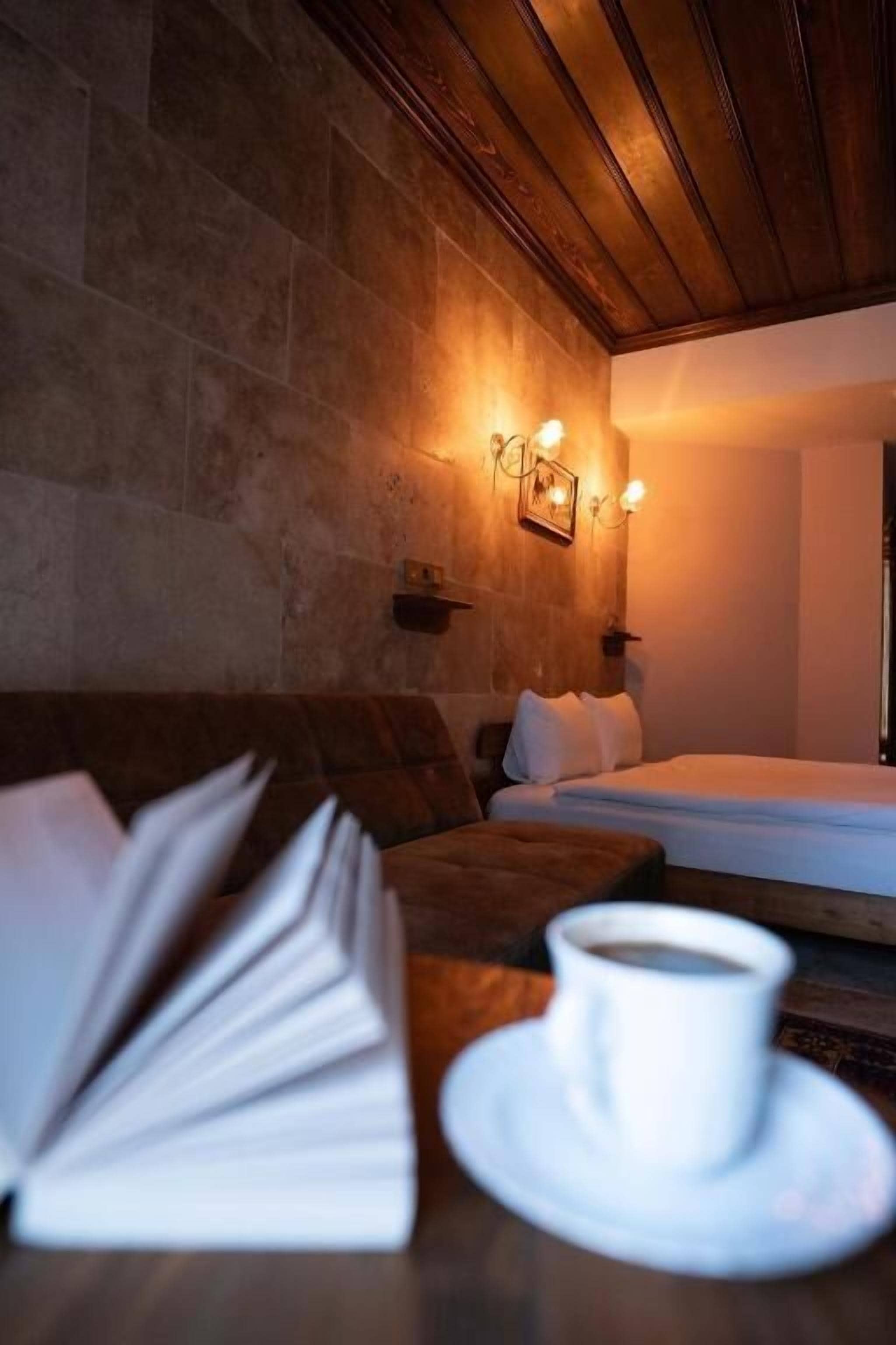 A cozy room with a stone wall, a wooden ceiling, a bed with white bedding, a brown sofa, and a table with a cup and saucer.