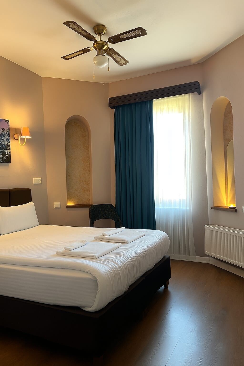 Hotellihuone, jossa on suuri sänky, kattotuuletin, sinisillä verhoilla varustettu ikkuna, seinään kiinnitetty lamppu ja patteri.
