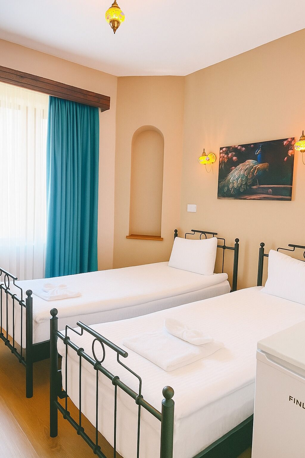 A hotel room with two single beds, a window with curtains, a painting on the wall, and a small shelf.