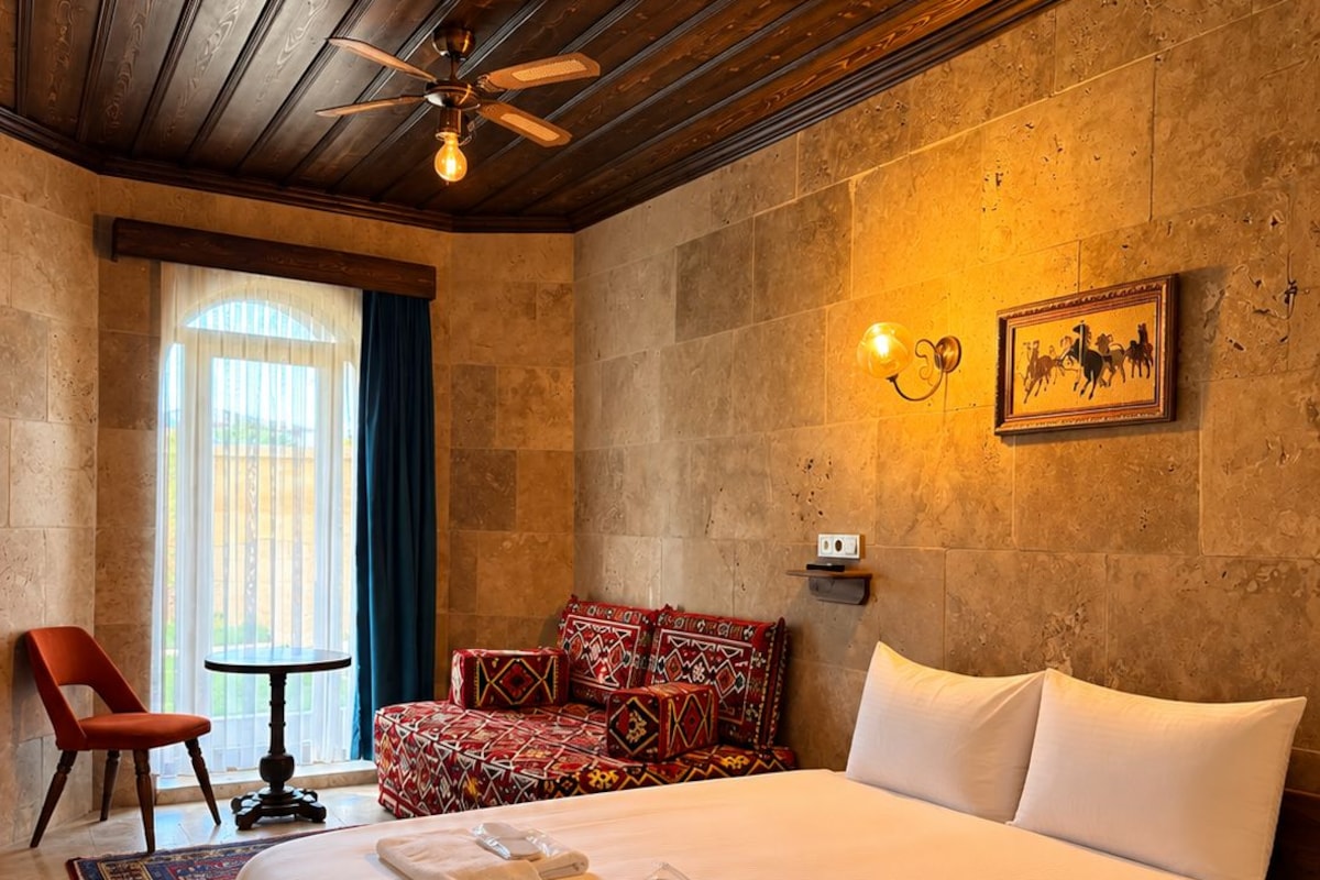 A hotel room with a large bed, a ceiling fan, a stone wall, a window with curtains, a red chair, and a small table.